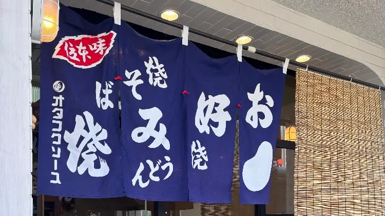 「お好み焼」「焼そば」と書かれた紺色ののれんが掛かるアサカナキッチン ルーブルーの店先